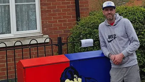 909 Art Mark James with short hair and beard leaning on a black and red painted cabinet