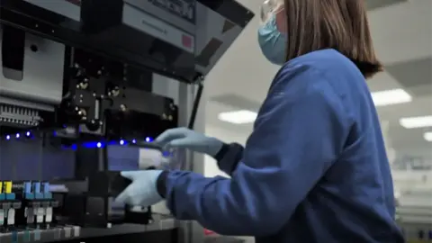 BBC/Brinkworth Productions Ltd A person in a face covering and latex gloves handles DNA samples in a laboratory