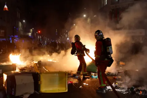 GONZALO FUENTES/Reuters Firefighters extinguish a fire during a demonstration, after the pension reform was adopted as the French Parliament rejected two motions of no-confidence against the government, in Paris, France, March 21, 2023.