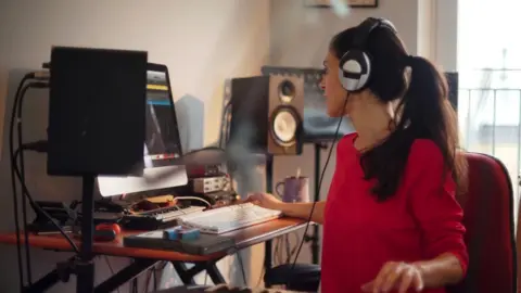 Getty Images A woman with headphones on and surrounded by music equipment sits at her computer