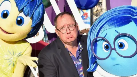 Getty Images John Lasseter is pictured comically frowning, seated between the characters of Joy and Sadness from Pixar's Inside Out in 2015