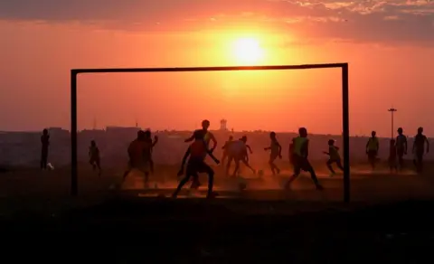 Reuters People are seen playing football through the goalposts. The sun appears large and hazy in the sky.