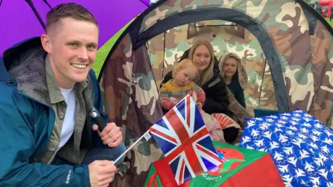 BBC Mum and two children cuddling inside a camouflage tent with dad crouched outside waving a Union flag