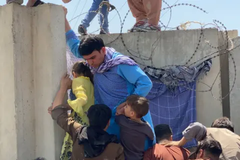 EPA People struggle to cross the boundary wall of Hamid Karzai International Airport to flee Afghanistan on 16 August 2021