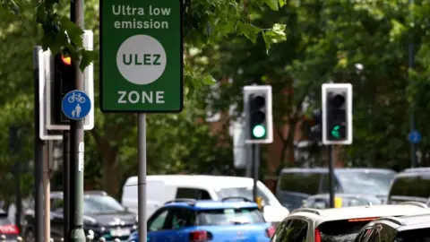 Reuters File image of stationary cars at traffic lights below a green Ulez sign.