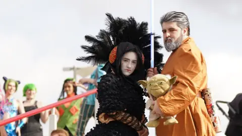 PA Media A woman dressed as Padme Amidala and a man dressed as Luke Skywalker holding a Yoda toy