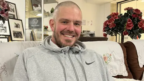 Jon Wright/BBC A man sat on a sofa, smiling