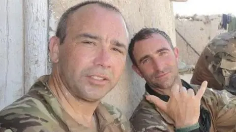 Facebook: Mark Townsend Two men in military uniforms pose for the camera, leaning against a wall in an arid location.