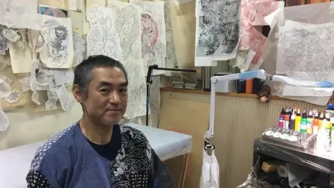 BBC Tattoo artist Horimitsu sitting in his studio in Tokyo, where the walls are lined with tattoo sketches