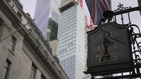 BBC An ornate green and gold sign for Simpson's hangs in front of a backdrop of skyscrapers in the city of London.