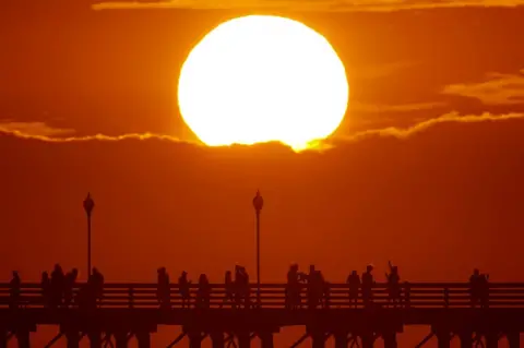 Mike Blake/Reuters The sun sets behind a cloud as people cool off with a walk along an ocean pier in Oceanside, California, US on 5 July 2018