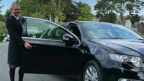 Ayesha Slader Ayesha Slader in funeral director's outfit holds car door open