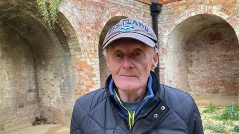 Man in late 70s wearing a blue cap with "Scotland" legend stands in front of brick arches.