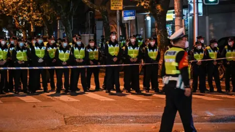 Getty Images Wearing fluorescent jackets and face masks, a row of police officers stand watch behind a cordon