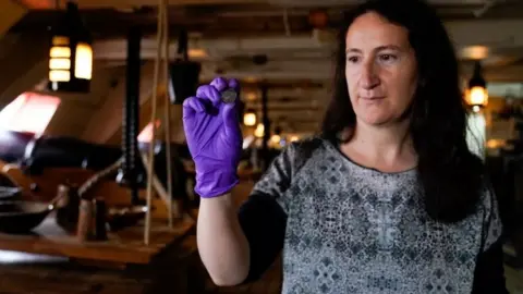 PA Media Diana Davis, Head of Conservation at the National Museum of the Royal Navy, holds a 127-year-old farthing on the gun deck of HMS Victory