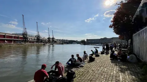 BBC People sitting on the harbour wall in Bristol with the MShed cranes visible in the background