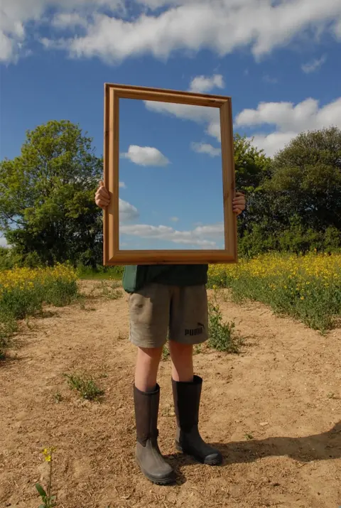 Lauren Sargeant A person stands in a field and holds a mirror showing the sky