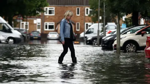 Carl Court/Getty Images A woman in wellies walks through floodwaters in a carpark near houses in Battersea, London