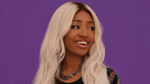 Jack Alexander A young, black woman with blonde hair stands in front of a deep purple plan background. She looks off to the side, smiling, her long hair spilling past her shoulders. She's wearing a chunky silver chain, both ends of are linked together by two fairly large hoops than join just under her throat.