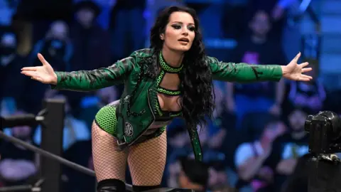 AEW Saraya stands on the turnbuckle of the wrestling ring, wearing a green leather jacket. Her arms are outstretched as she interacts with the crowd