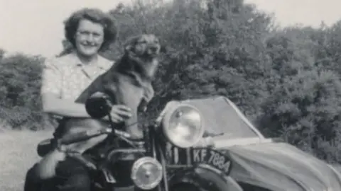 Family handout Old black and white photo of Pauline Polhill. She is smiling at the camera whilst sitting on a motorcycle next to a large dog