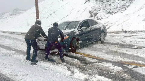 AFP via Getty Images Two men try to stop a car from sliding down an icy roads during a snow storm in Rancho Cucamonga, California. Photo: 25 February 2023