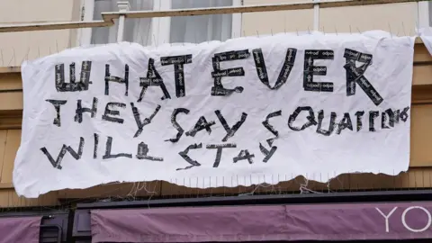 Lucy North Sign on bedsheet outside pub saying: Whatever they say squatters will stay