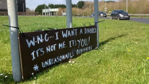 Martin Heath/BBC Black sign with white lettering on a roundabout. The sign says: "WNC... I want a divorce"