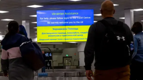 Getty Images People walk past an information billboard at London Luton Airport designed for Ukrainian refugees who arrive in the United Kingdom