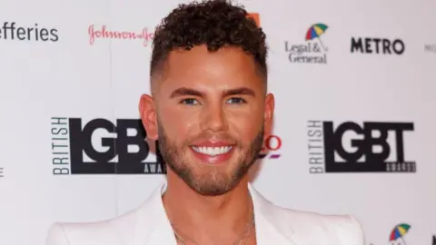 Getty Images Dean McCullough attends the British LGBT Awards 2023 in London. Dean is a 31-year-old white man with short curly brown hair on top of his head, with shaved sides. He has a stubbly brown beard and blue eyes. He wears a white blazer and smiles at the camera.