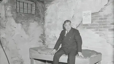 Bettmann/Getty Black and white photo showing a middle-aged man dressed in suit and tie sitting on a basic bed in a room with brick and stripped off plaster and a small window with bars