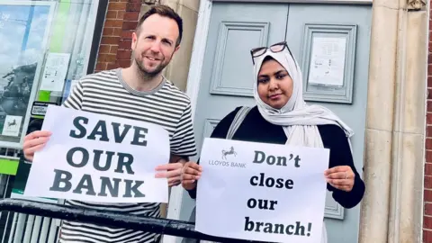 LDRS Councillors Ben Miskell and Nabeela Mowlana standing outside Lloyds Bank holding placards