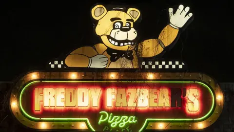 Patti Perret/Universal Pictures A slightly grubby neon sign that reads "Freddy Fazbear's Pizza" in block capital letters. Above the lettering is an illuminated bear character. He's smiling and has one arm raised in a wave.