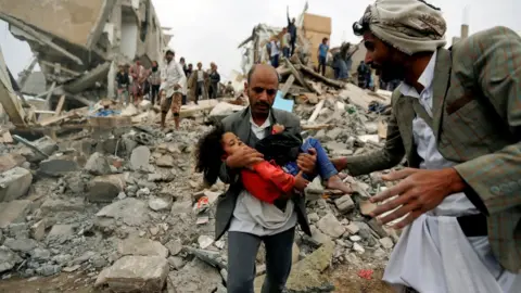 Reuters A man carries Buthaina Muhammad Mansour from the site a Saudi-led coalition air strike that killed members of her family in Sanaa, Yemen (25 August 2017)