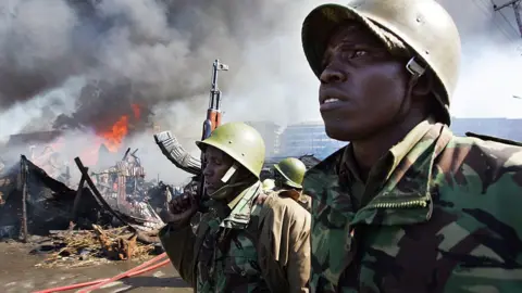 AFP Kenyan police walk past burning shacks in the Mathare Slum 30 December 2007 in Nairobi, after fresh violence linked to the disputed presidential poll erupted.