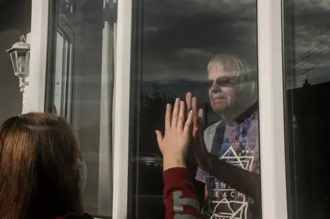 Jessica Shackleford A young person holds their hand up to the window as a relative looks on from the other side