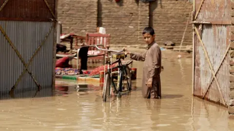 BBC A boy stands with his bicycle in knee-deep muddy water in Nowshera