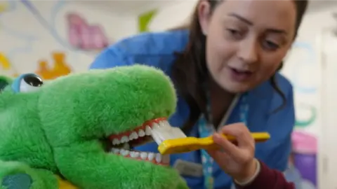 John Fairhall/BBC dental nurse and child brushing a dinosaur's teeth