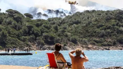 AFP/Getty Images People sit on the beach as they look at Canadair aircraft dropping water over a fire in La Croix-Valmer, near Saint-Tropez. Photo: 25 July 2017
