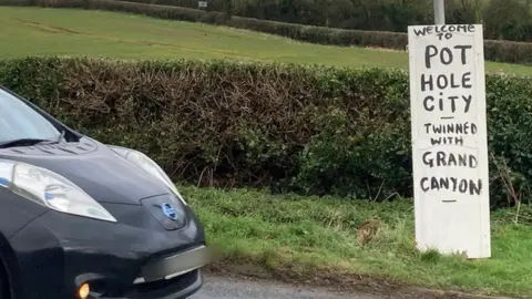 Martin Heath/BBC Sign on rural road with black lettering welcoming drivers to Pot Hole City twinned with Grand Canyon.