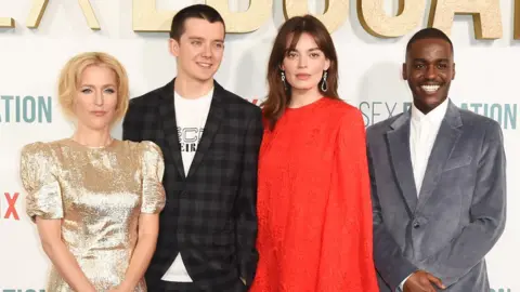 Getty Images Gillian Anderson, Asa Butterfield, Emma Mackey and Ncuti Gatwa attend the World Premiere of Netflix's "Sex Education" Season 2 at The Genesis Cinema on January 8, 2020 in London. Gillian, a blonde woman in her 40s, wears a gold short sleeved dress. Asa, on her left, is a white man in his 20s with very short dark hair - he wears a checked grey and black blazer over a white T-shirt. Next to him stands Emma, a white woman in her 20s with dark hair worn loose just past her shoulders. She is wearing a long sleeved, high-necked floaty red dress. On the end, Ncuti is a black man in his 20s wearing a grey suede blazer over a white shirt and smiling at the camera.