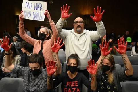 AFP Protesters raise red painted hands as US Secretary of State Antony Blinken and Defense Secretary Lloyd Austin testify during a Senate Appropriations Committee hearing on a funding request for Israel