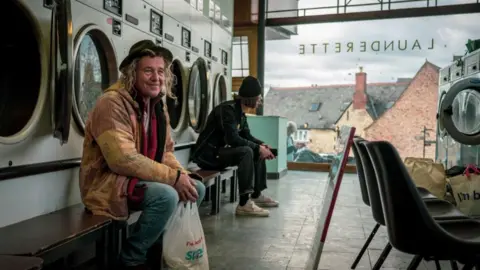 Rupert Russel A man sits in a launderette and smiles past the camera