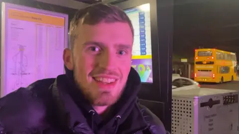 Andrew Turner/BBC Kyle Thompson stands at a bus stop in Norwich. Bus in background of picture