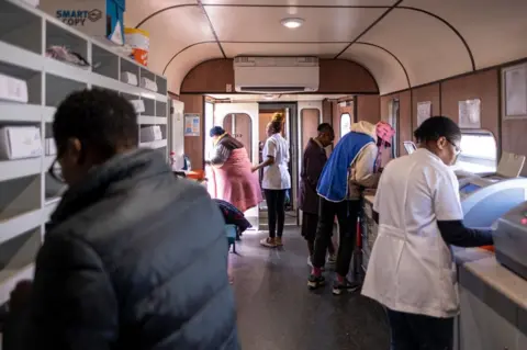 EMMANUEL CROSET/AFP Ophthalmic laboratory technicians prepare frames and lenses for the following patients as one climbs off the wagon of the Phelophepa train, parked at the Dube train station in Soweto, on October 17, 2023. The Transnet-Phelophepa trains are free, mobile healthcare clinics that travel to areas of rural South Africa, where there is just one doctor for every 5,000 patients. Phelophepa, a Sotho and Venda word meaning 'good, clean health', is owned and operated by Transnet Foundation.