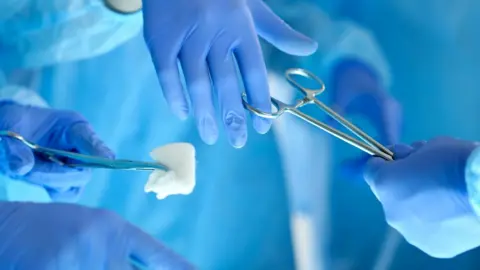 Blue-gloved hands of medical latex exchange a surgical scissors, while a third hand can be seen holding a clamp and cotton swab
