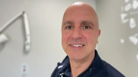 Dr Jason Lester in a clinical consulting room. He is wearing a navy top.