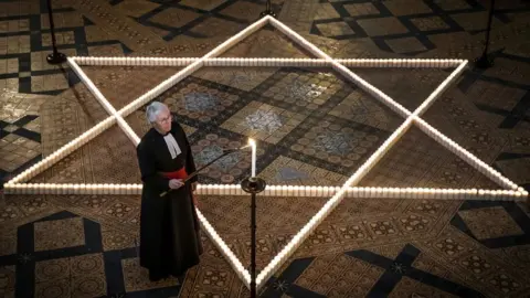 PA Media A reverend lights a candle on a stand. On a tiled floor behind him, hundreds of candles are arranged in a star