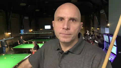 John Devine/BBC Joe Perry stood with a cue and snooker tables in the background