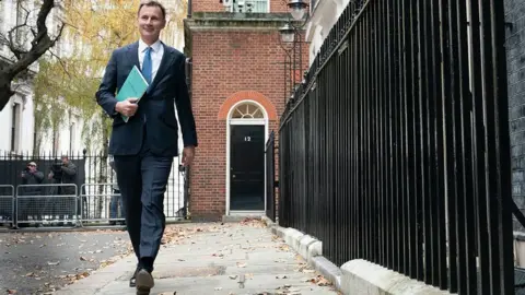 PA Media Jeremy Hunt walking outside number 11 Downing Street in a suit and blue tie holding a green budget statement in November 2023